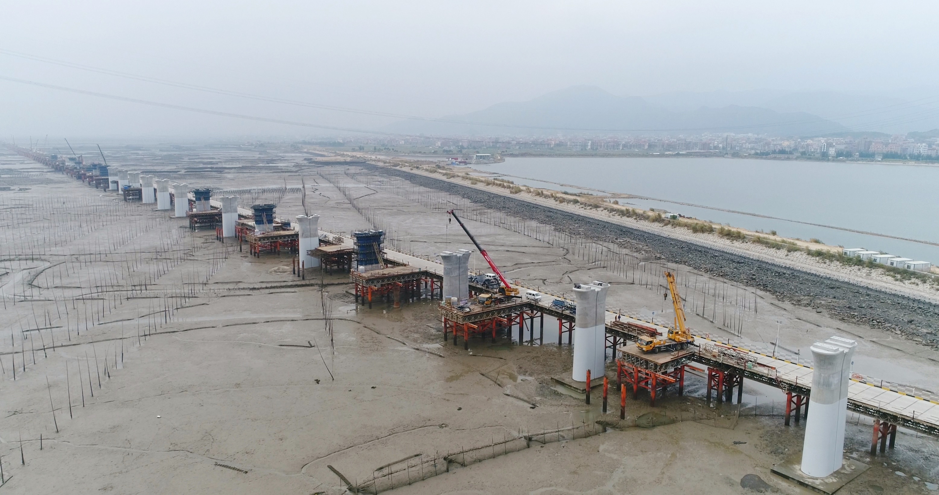 Long steel bailey bridge under construction over a muddy area with many pilings, extending towards a distant city, demonstrating high load capacity for heavy infrastructure projects